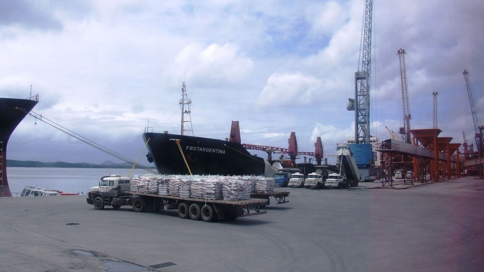 Carreta carregada de sacas brancas no cais do porto, ao lado de um navio preto chamado Frotargentina e de guindastes