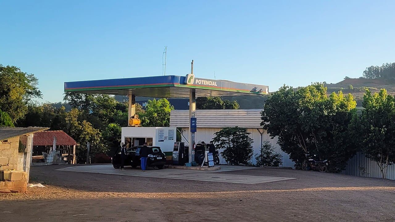 Posto de combustível pequeno em área rural, com cobertura azul, um carro abastecendo, galpão ao fundo e morros com vegetação