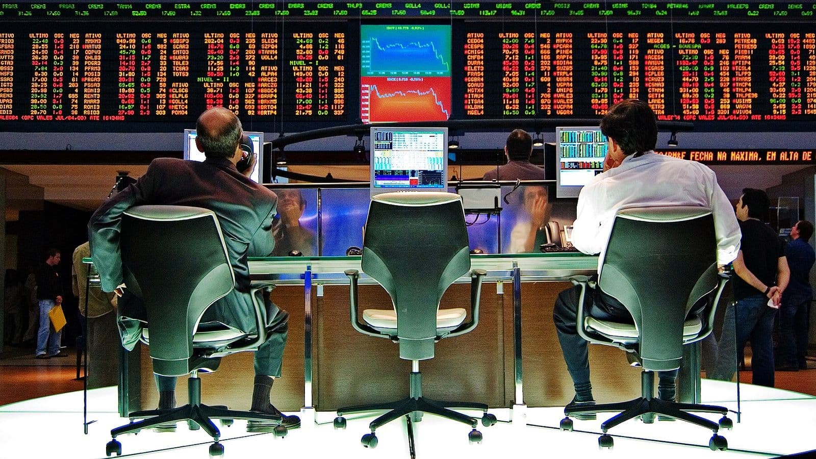 Dois operadores sentados de costas diante de monitores no pregão da Bovespa, com um painel eletrônico de cotações em vermelho e verde ao fundo