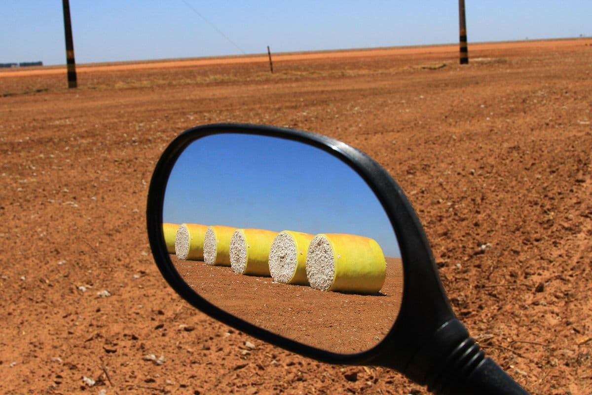 Retrovisor de moto reflete uma fila de rolos de algodão colhido, embalados em plástico amarelo, sobre terra vermelha sob céu azul