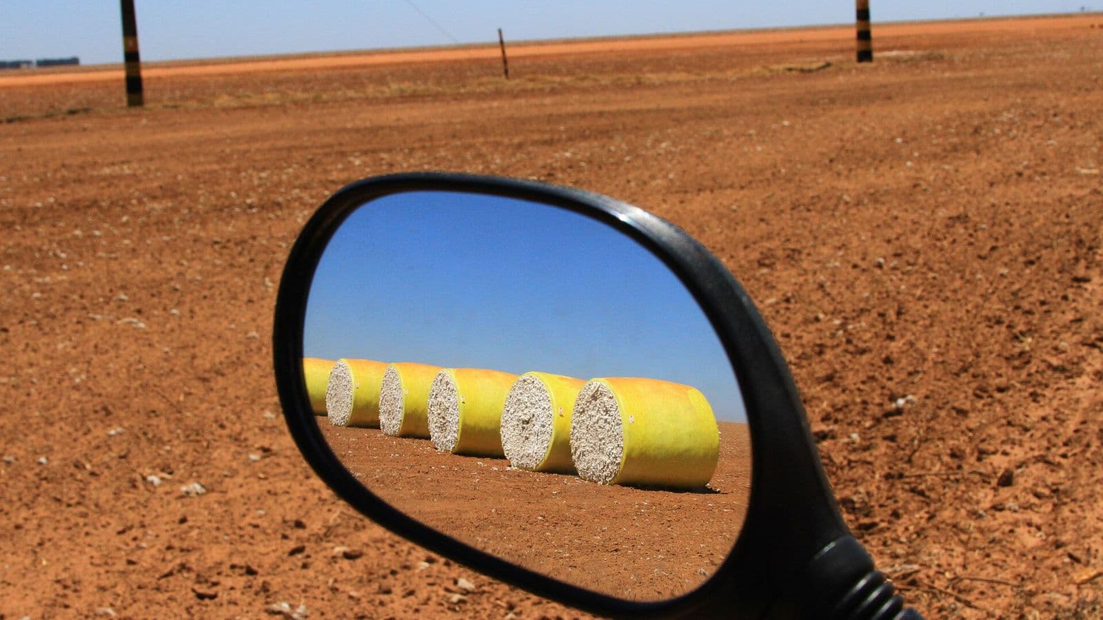 Retrovisor de moto reflete uma fila de rolos de algodão colhido, embalados em plástico amarelo, sobre terra vermelha sob céu azul