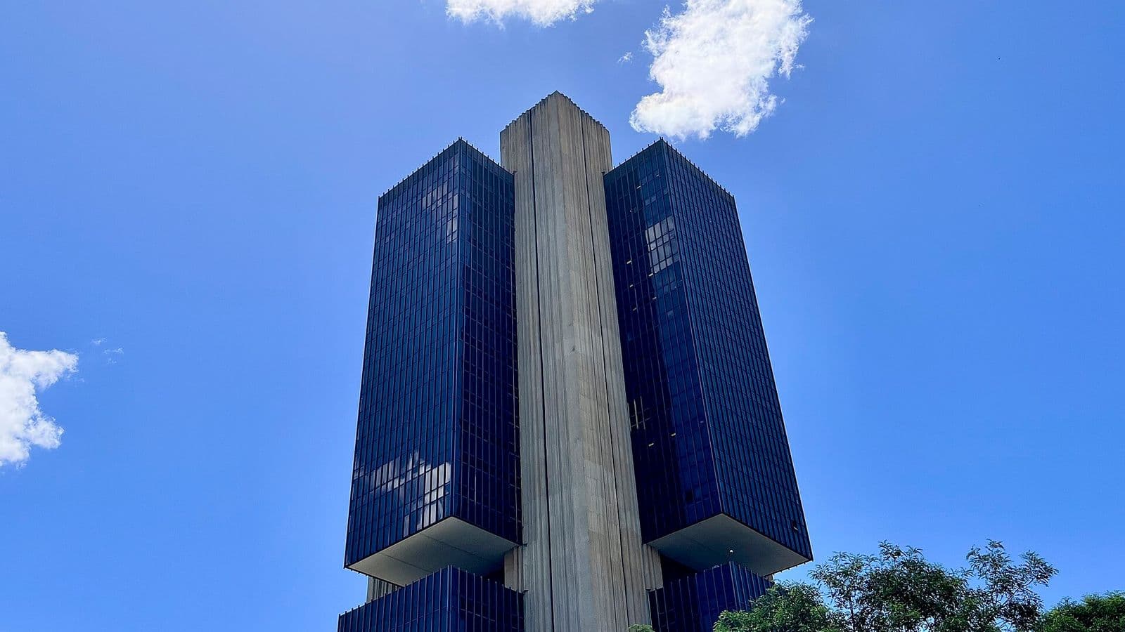 Edifício-sede do Banco Central em Brasília, torre escura de vidro com pilar central de concreto, vista de baixo contra o céu azul, com árvores em primeiro plano.
