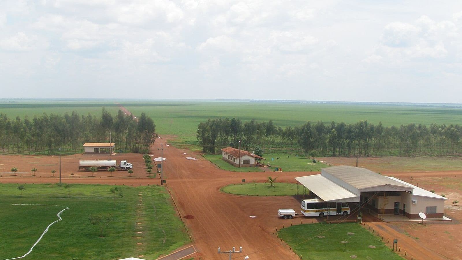 Vista do alto da sede de uma fazenda, com galpões, estradas de terra vermelha, fileiras de eucalipto e lavouras verdes até o horizonte