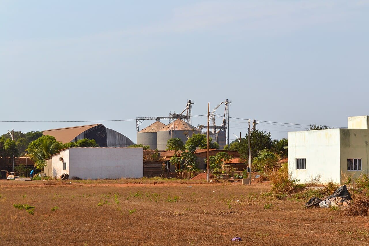 Silos metálicos de armazenagem de grãos com torres de elevação, atrás de casas e de um galpão de cobertura arredondada, em terreno de terra seca