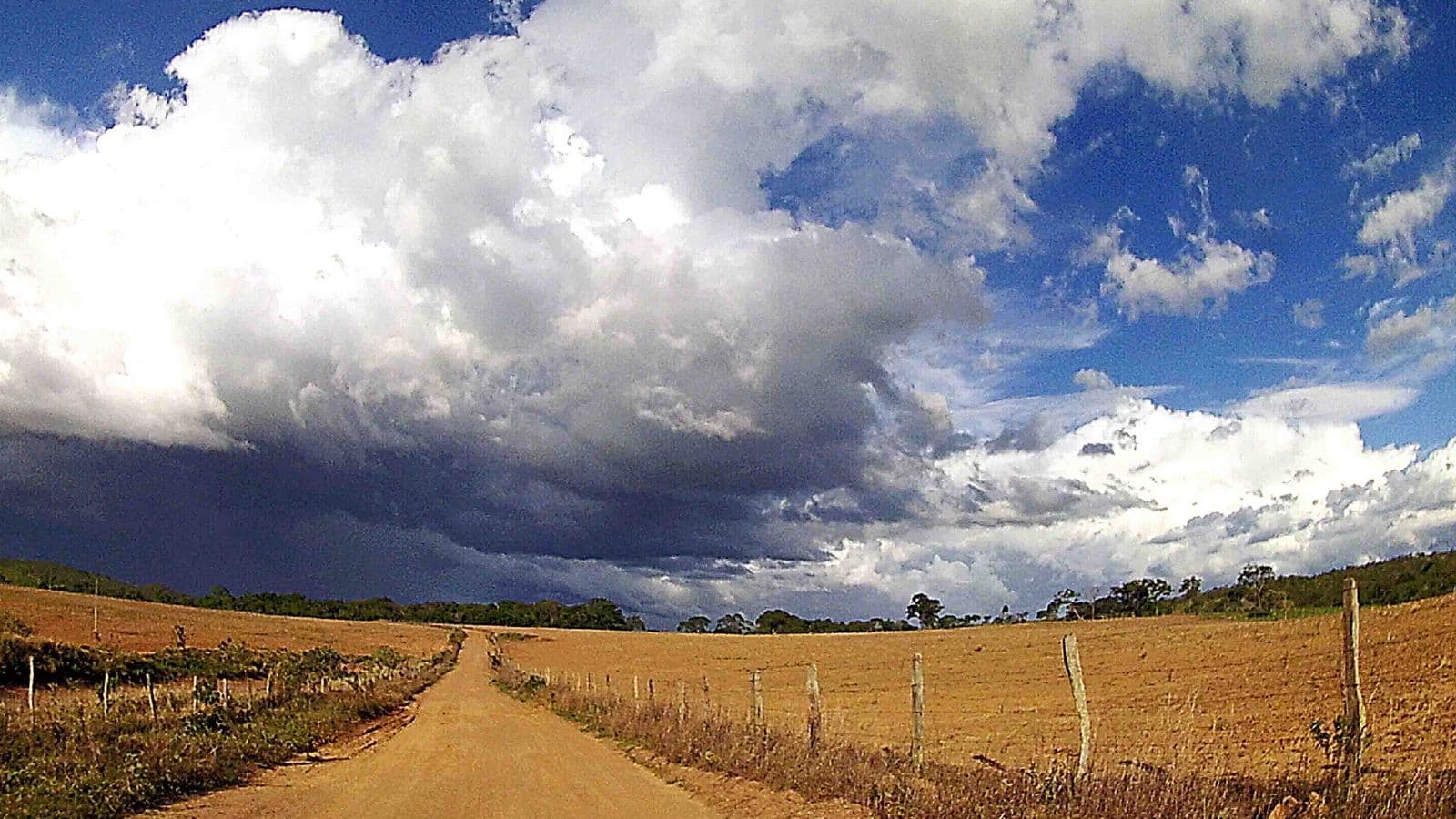 Estrada de terra entre pastos secos sob nuvens escuras de tempestade e céu azul em área rural de Goiás