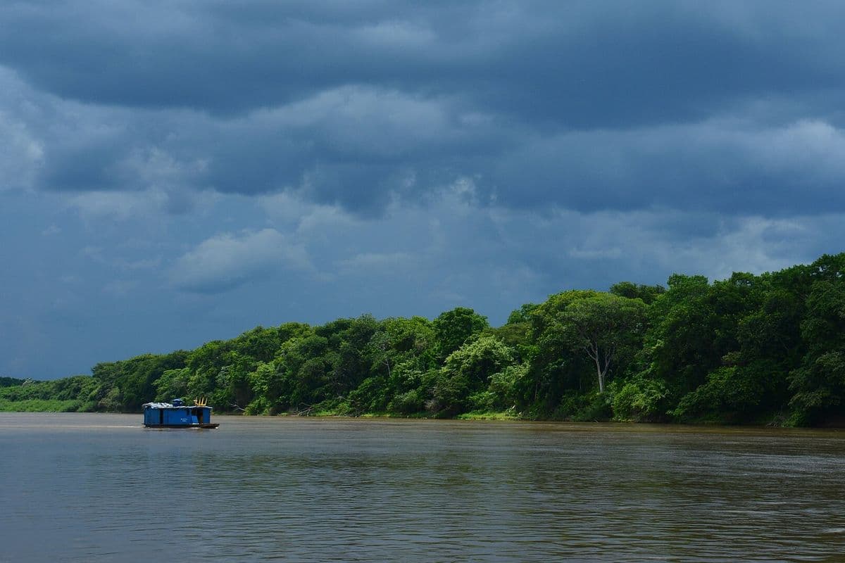 Nuvens escuras de tempestade cobrem o céu sobre um rio largo de água barrenta, com mata densa na margem e um pequeno barco azul