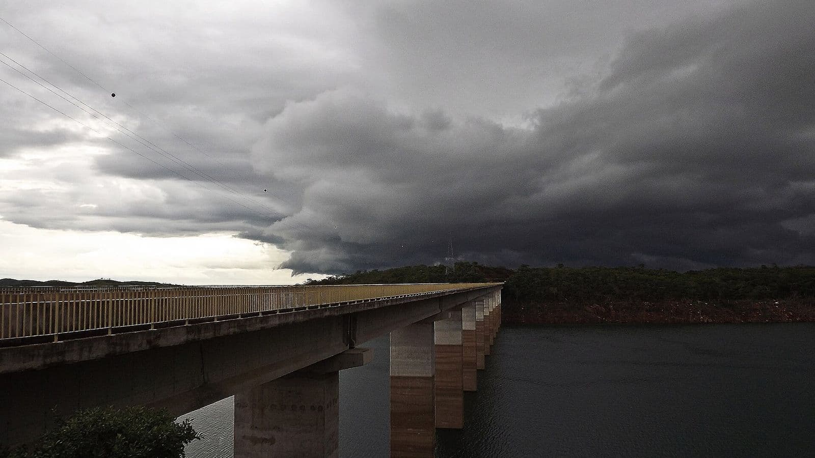 Nuvens escuras e baixas de tempestade avançam sobre um lago, com uma longa ponte de concreto à esquerda e mata na margem ao fundo