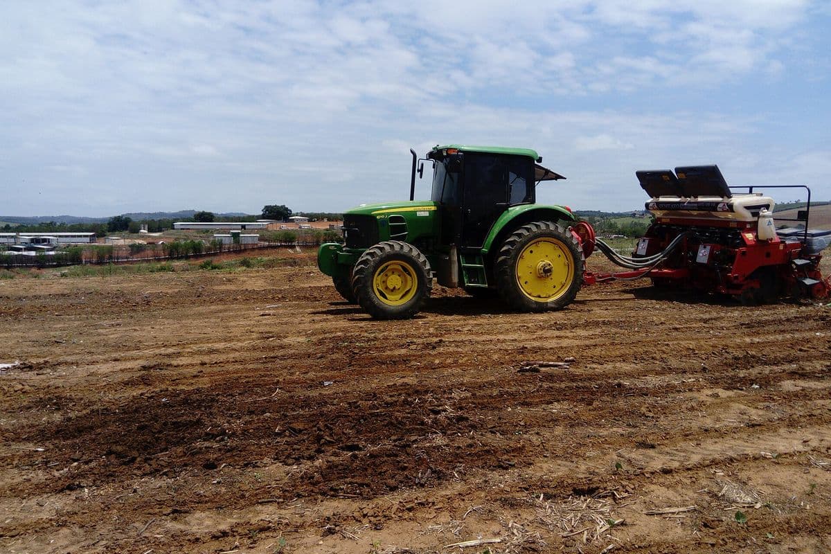 Trator verde puxa plantadeira vermelha sobre terra preparada, com galpões e céu azul com nuvens ao fundo