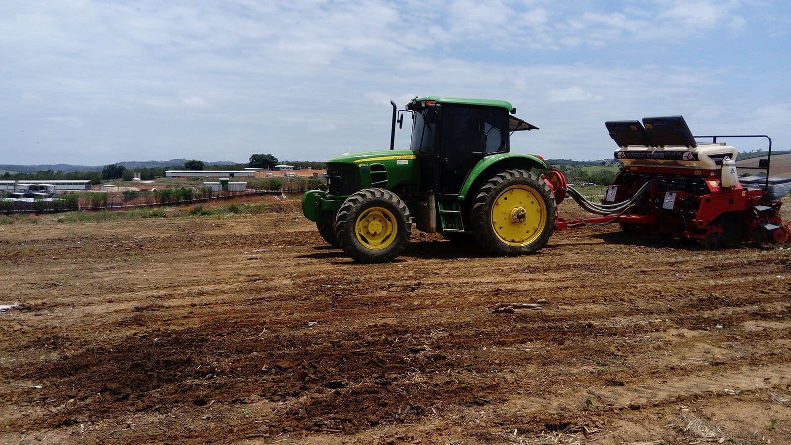 Trator verde puxa plantadeira vermelha sobre terra preparada, com galpões e céu azul com nuvens ao fundo