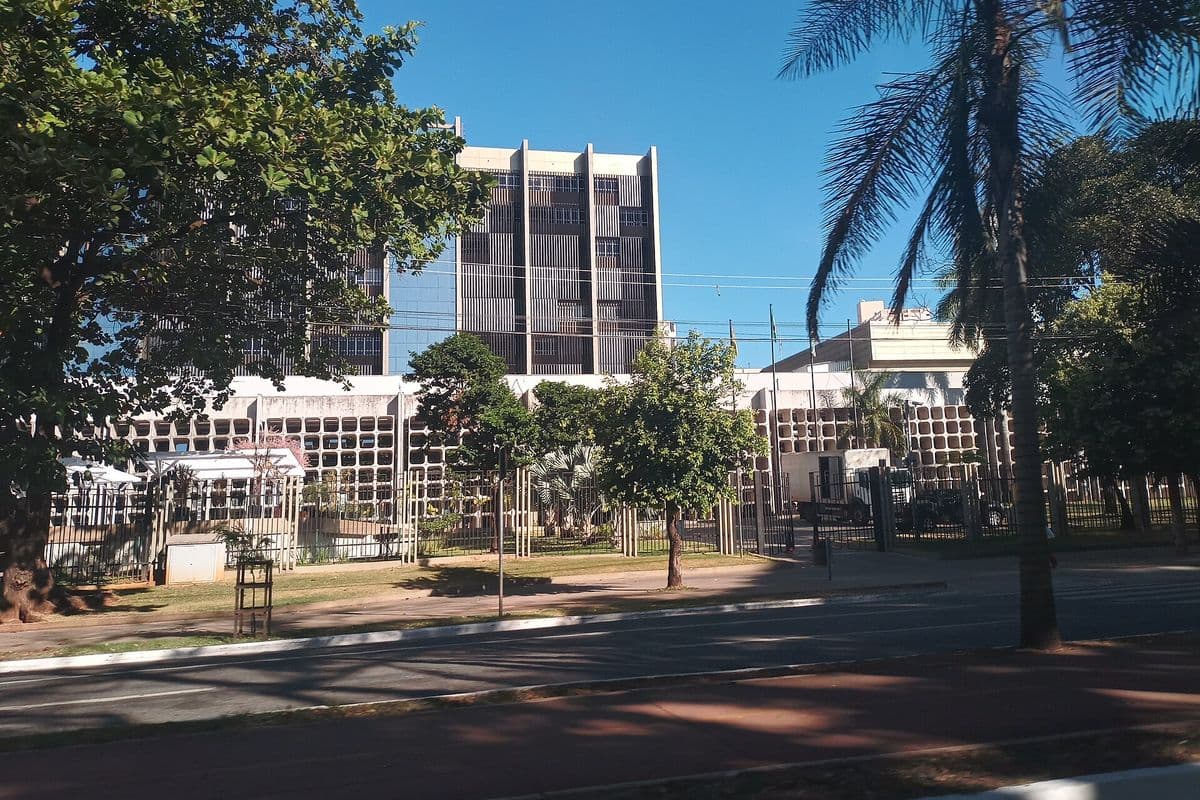 Prédio do Tribunal de Justiça de Goiás, com uma torre alta de fachada marrom e um bloco baixo branco à frente, cercado por grades e árvores, visto do outro lado de uma avenida sob céu azul