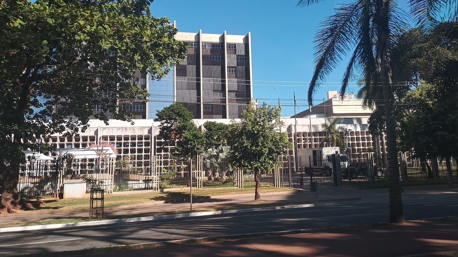 Prédio do Tribunal de Justiça de Goiás, com uma torre alta de fachada marrom e um bloco baixo branco à frente, cercado por grades e árvores, visto do outro lado de uma avenida sob céu azul