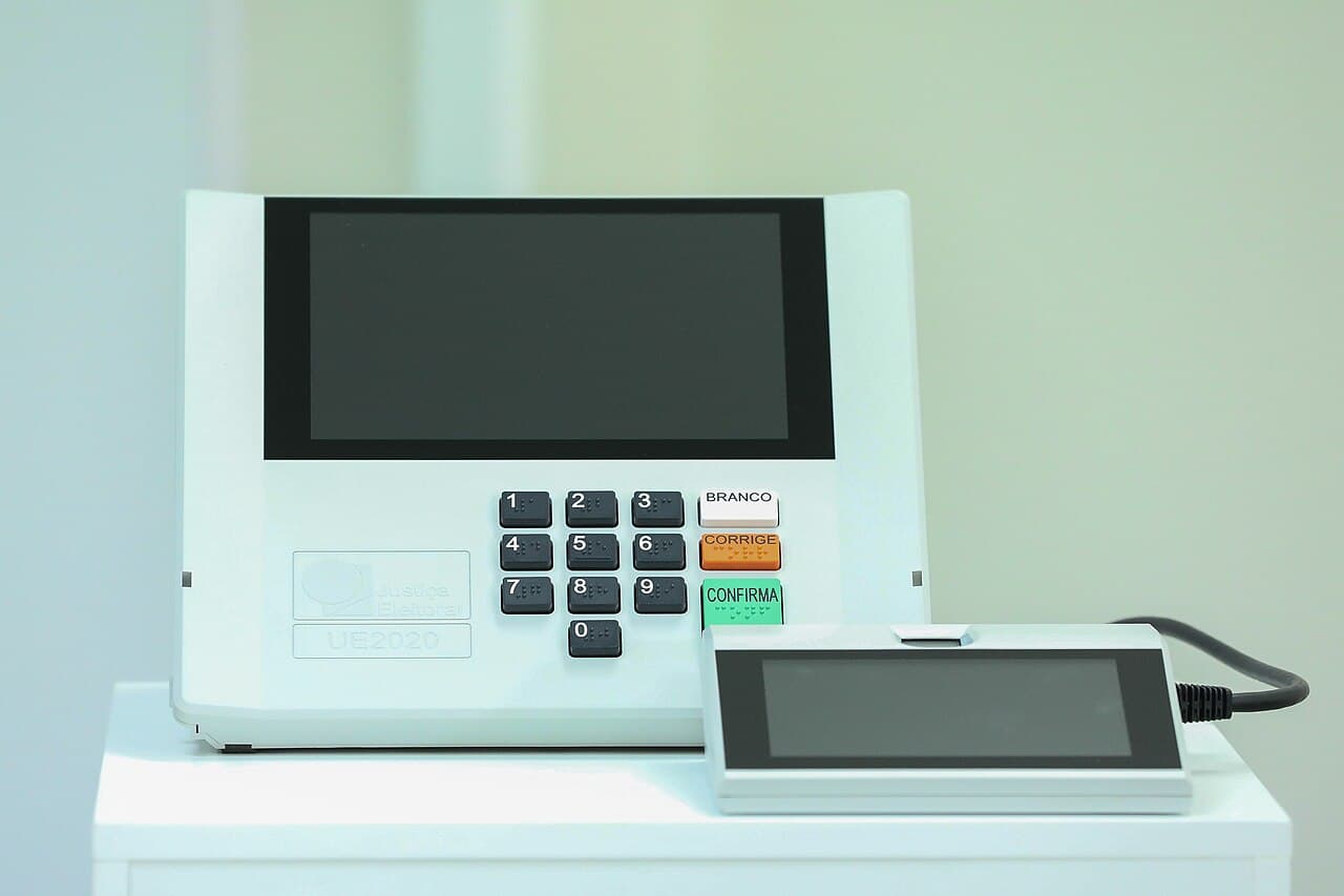 Urna eletrônica brasileira modelo UE2020, com tela, teclado numérico e as teclas branco, corrige e confirma, ao lado do terminal do mesário