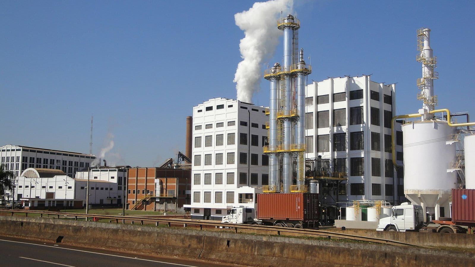 Usina com colunas metálicas de destilação soltando vapor branco, prédios industriais brancos, tanques e caminhões com contêineres diante de uma rodovia, sob céu azul