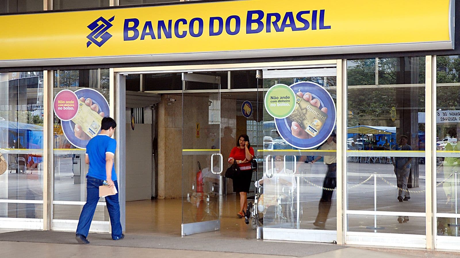 Fachada de agência do Banco do Brasil com letreiro amarelo e portas de vidro, com um homem entrando e uma mulher saindo da agência