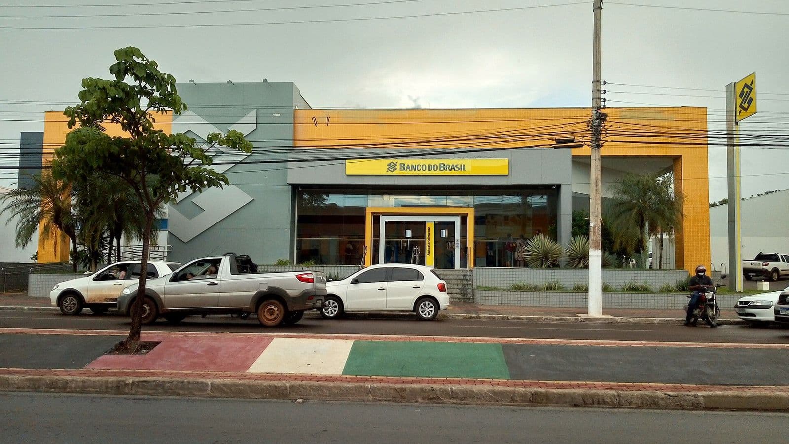 Fachada amarela e cinza de agência do Banco do Brasil, com carros estacionados em frente, poste com fios e céu nublado