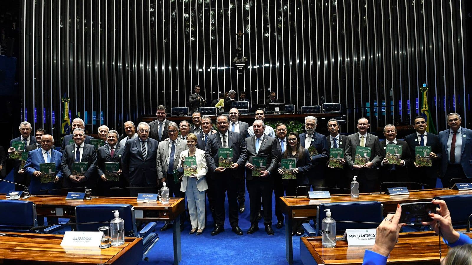 Cerca de 30 parlamentares e dirigentes, em trajes sociais, posam em fileiras no plenário segurando exemplares de capa verde de um documento; ao fundo, painel prateado de lâminas verticais e a mesa diretora.