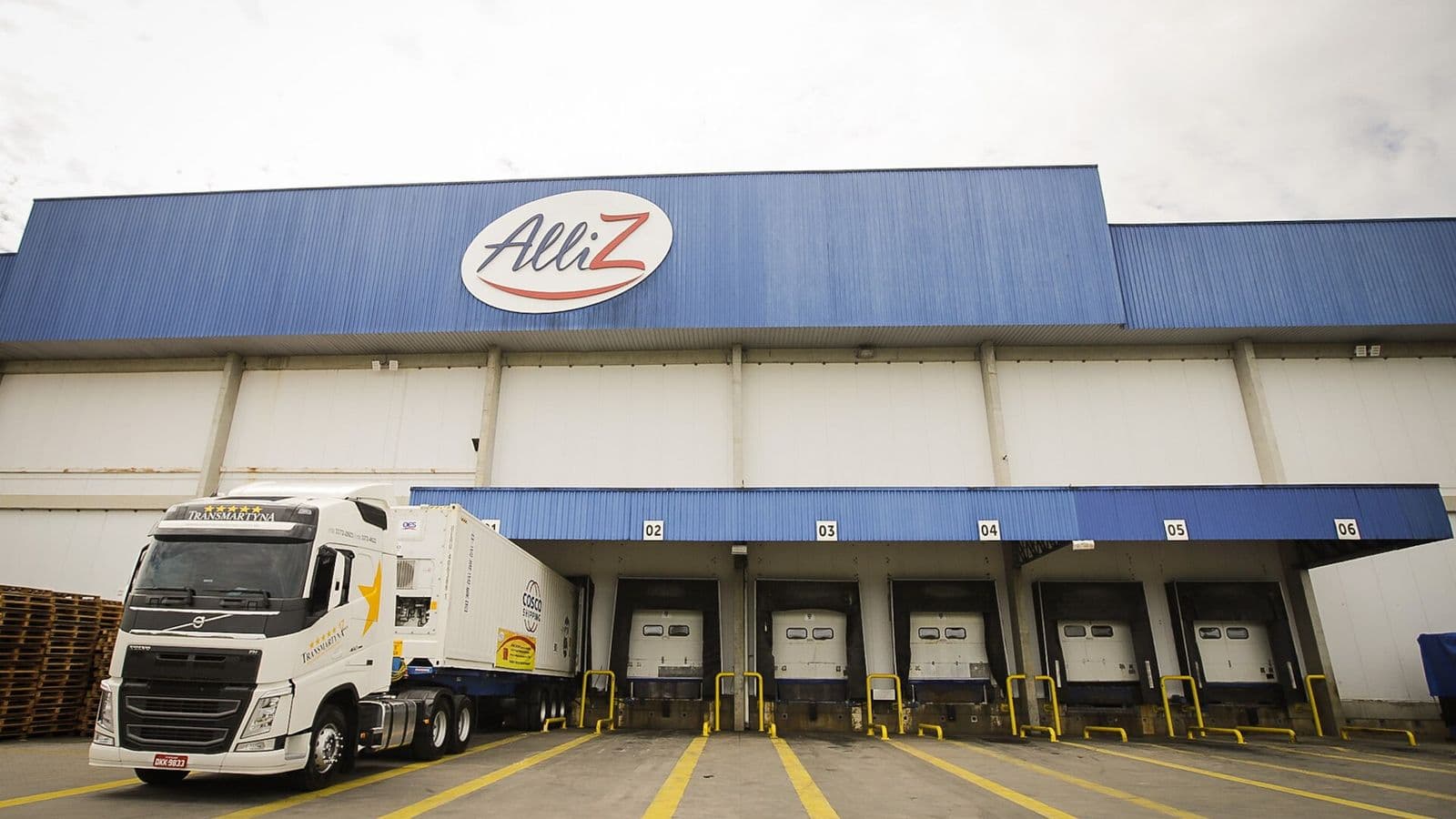 Caminhão branco com contêiner refrigerado encostado nas docas de um armazém frigorífico branco e azul, sob céu nublado