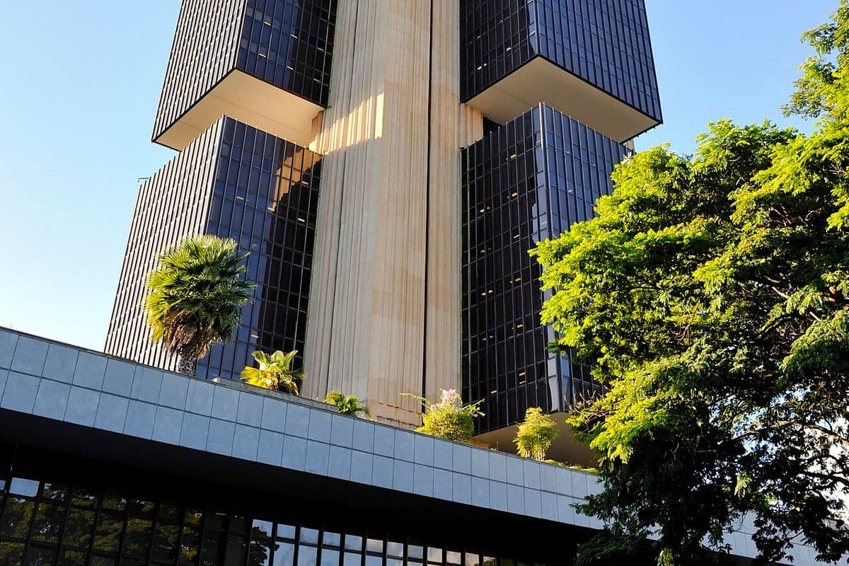 Torre de vidro escuro e concreto do edifício-sede do Banco Central, em Brasília, com palmeiras e uma árvore em primeiro plano