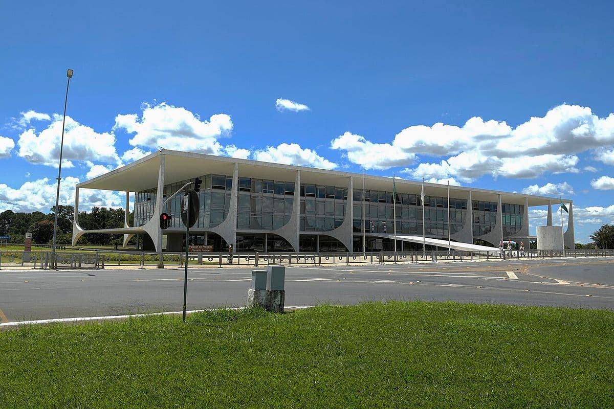 Fachada envidraçada do Palácio do Planalto, com suas colunas curvas brancas, vista do outro lado da avenida, sob céu azul com nuvens