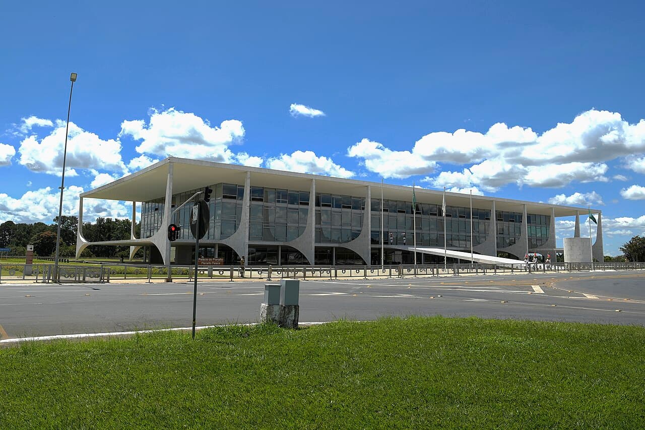 Fachada envidraçada do Palácio do Planalto, com suas colunas curvas brancas, vista do outro lado da avenida, sob céu azul com nuvens