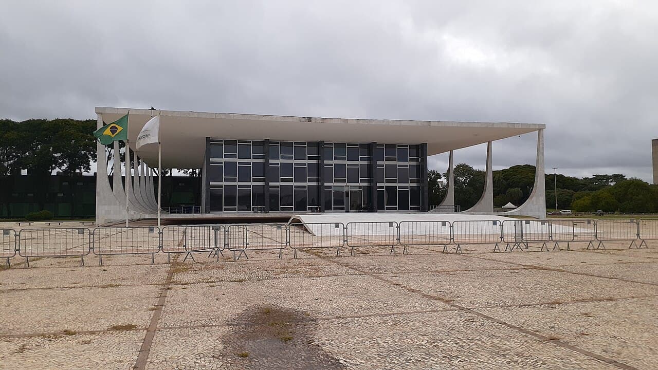 Fachada do Palácio do Supremo Tribunal Federal, com colunas curvas brancas, bandeira do Brasil e grades na praça em frente, sob céu nublado