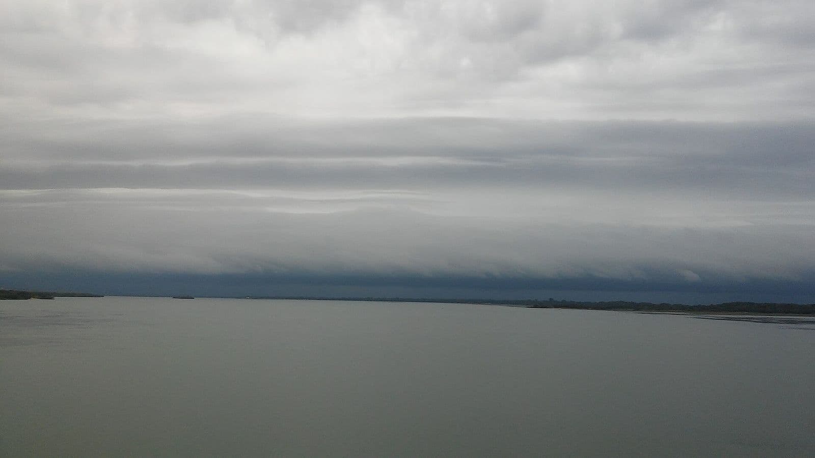 Faixa espessa de nuvens cinza-escuras, em camadas horizontais, avança no horizonte sobre a superfície larga e calma de um rio, sob céu encoberto
