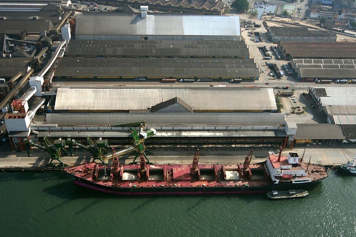 Vista aérea de um navio graneleiro com porões abertos atracado diante de armazéns e esteiras de um terminal de grãos