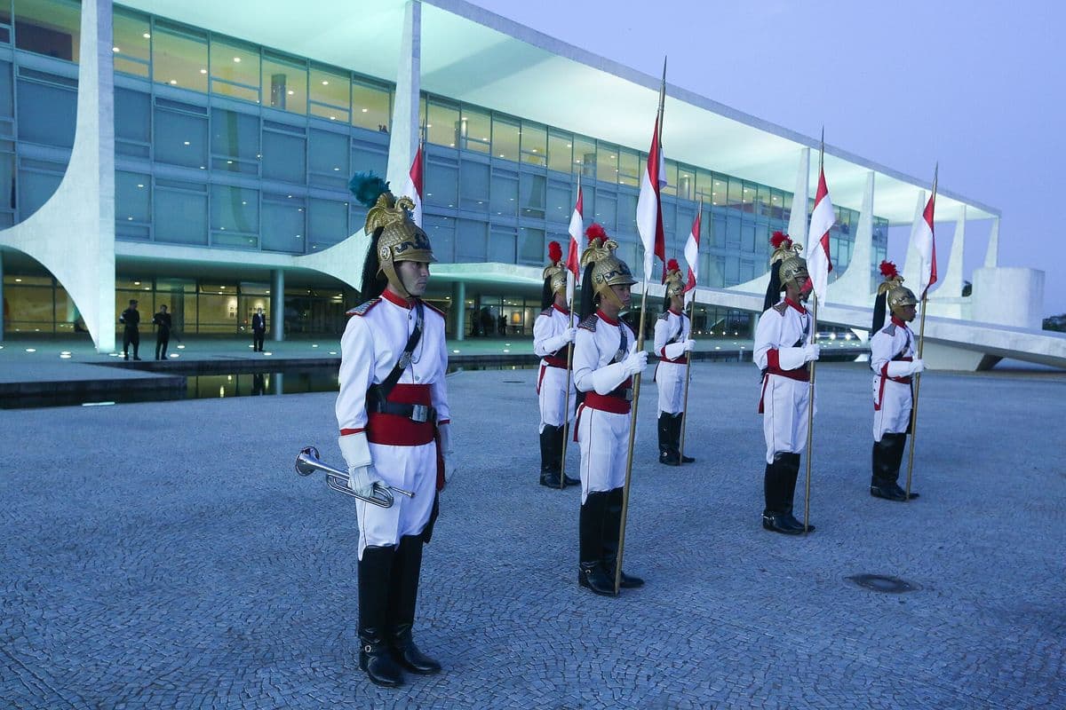 Cinco soldados dos Dragões da Independência, de uniforme branco e vermelho e capacete dourado com penacho, perfilados com lanças diante da fachada iluminada do Palácio do Planalto ao anoitecer