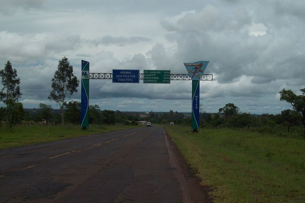 Rodovia de asfalto passa sob pórtico azul e verde com placas de boas-vindas e de distâncias, sob céu carregado de nuvens