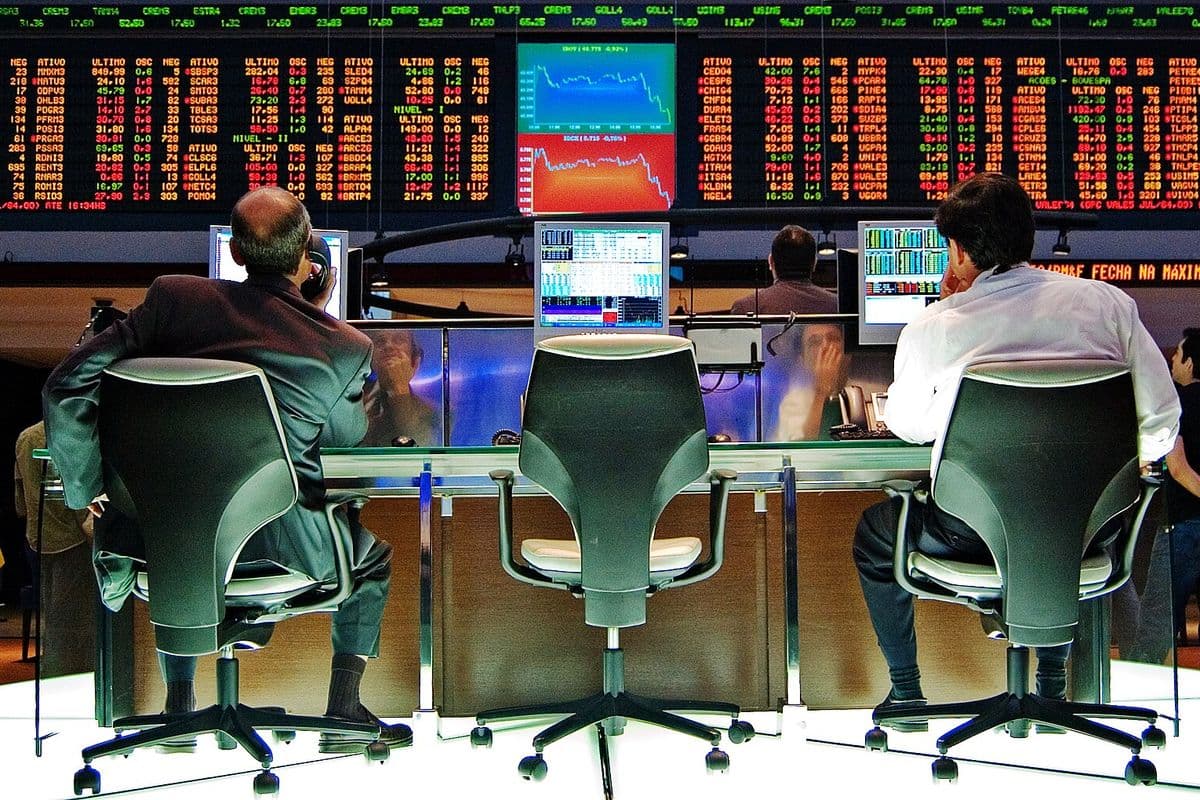 Dois operadores sentados de costas diante de monitores no pregão da Bovespa, com um painel eletrônico de cotações em vermelho e verde ao fundo