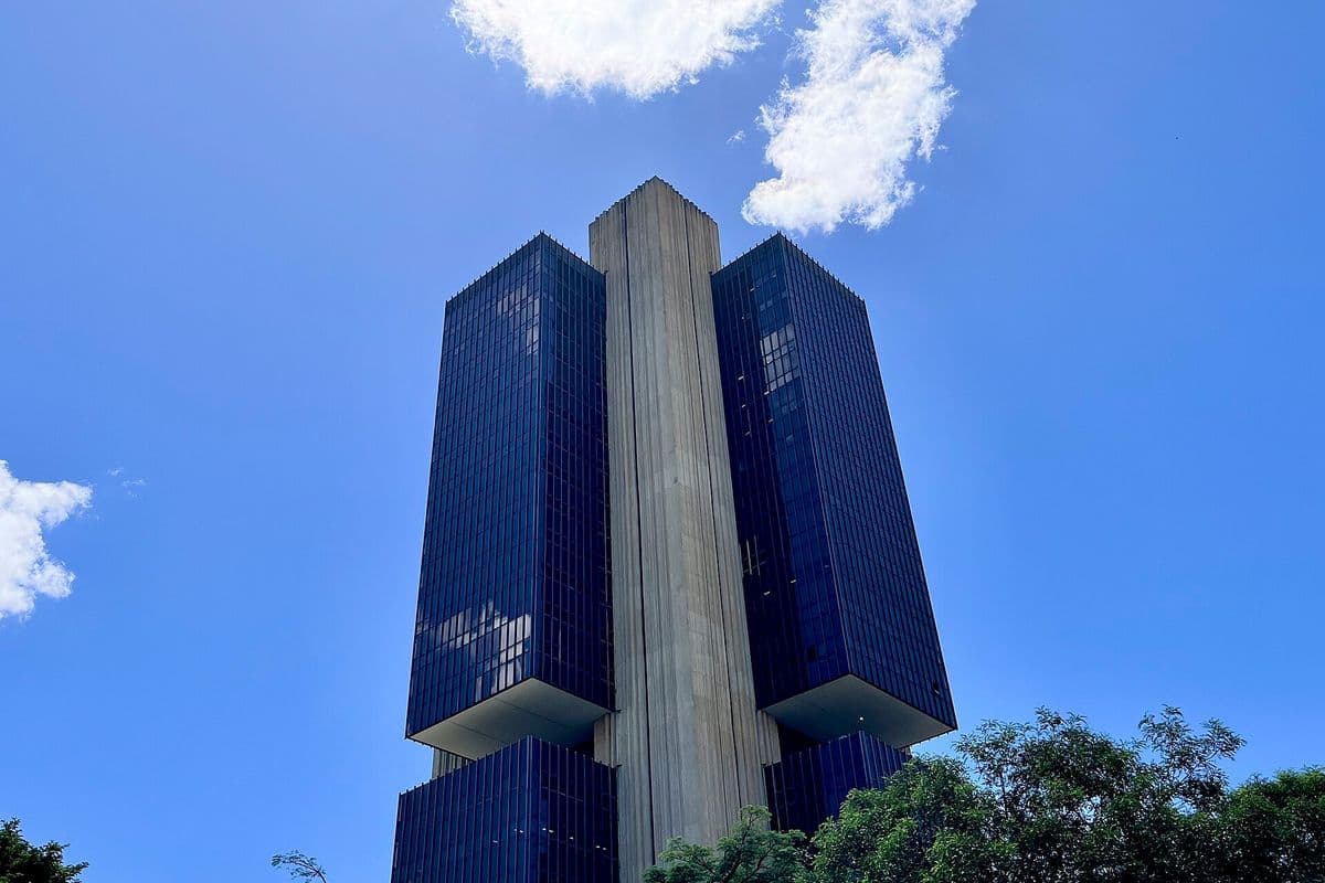 Edifício-sede do Banco Central em Brasília, torre escura de vidro com pilar central de concreto, vista de baixo contra o céu azul, com árvores em primeiro plano.