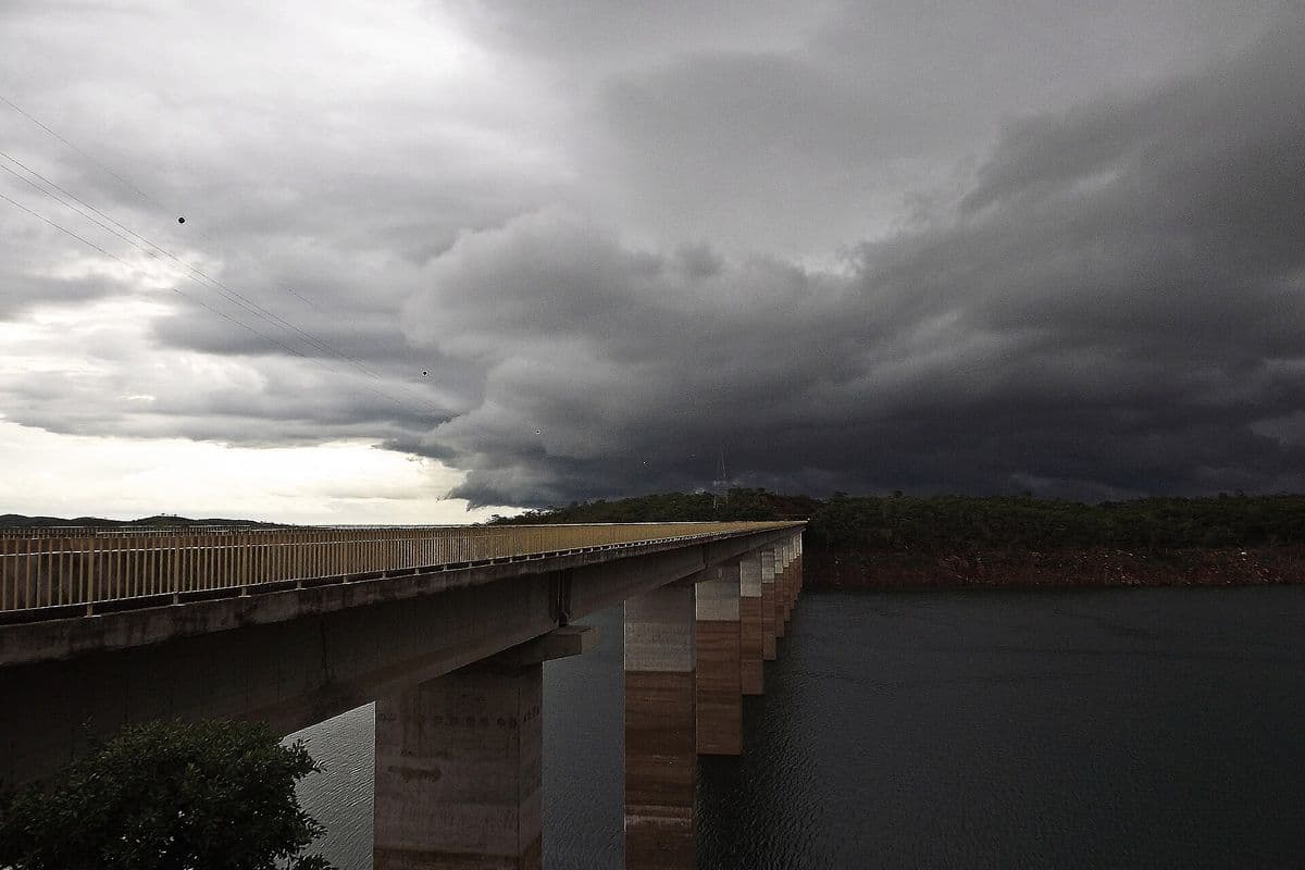 Nuvens escuras e baixas de tempestade avançam sobre um lago, com uma longa ponte de concreto à esquerda e mata na margem ao fundo