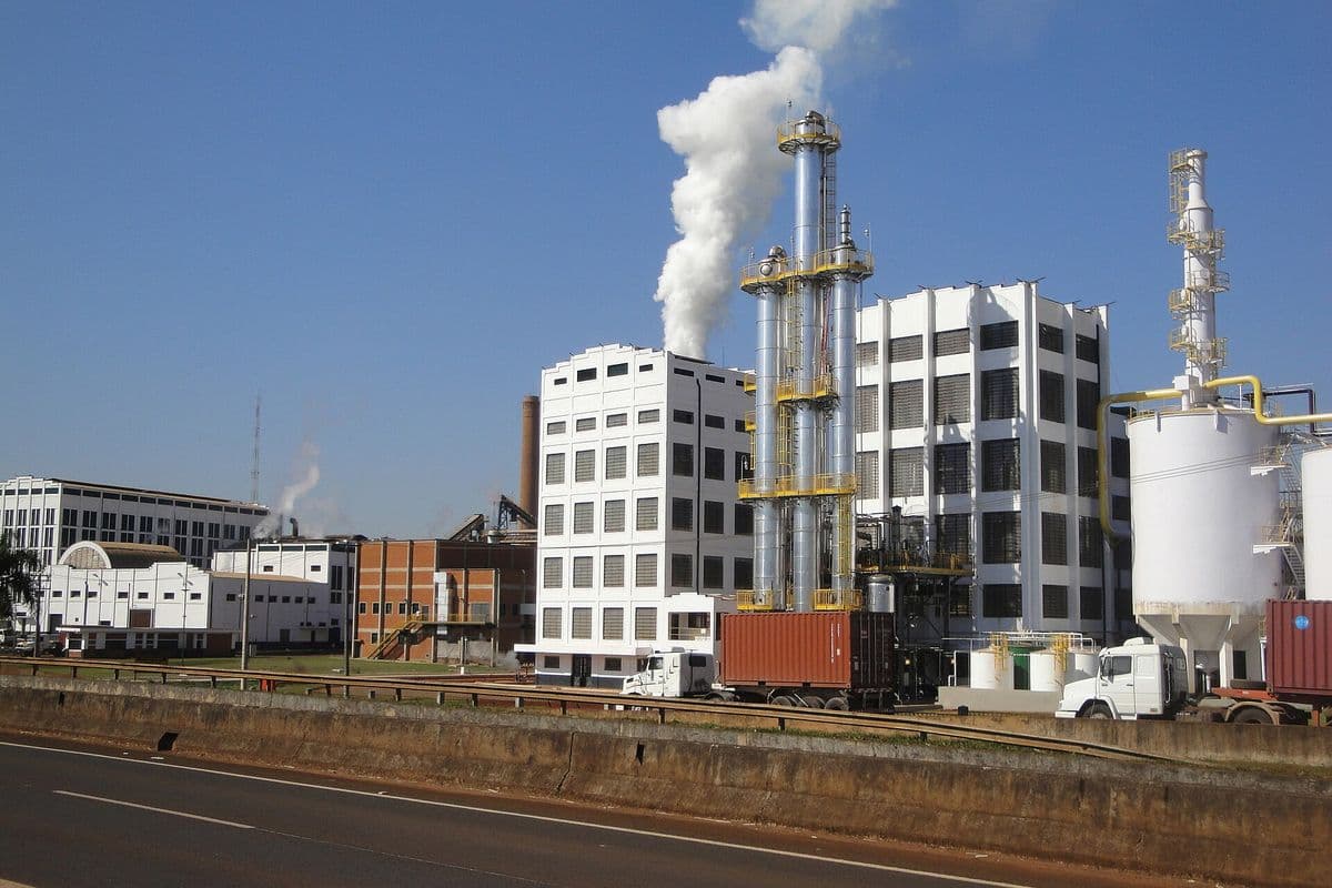 Usina com colunas metálicas de destilação soltando vapor branco, prédios industriais brancos, tanques e caminhões com contêineres diante de uma rodovia, sob céu azul